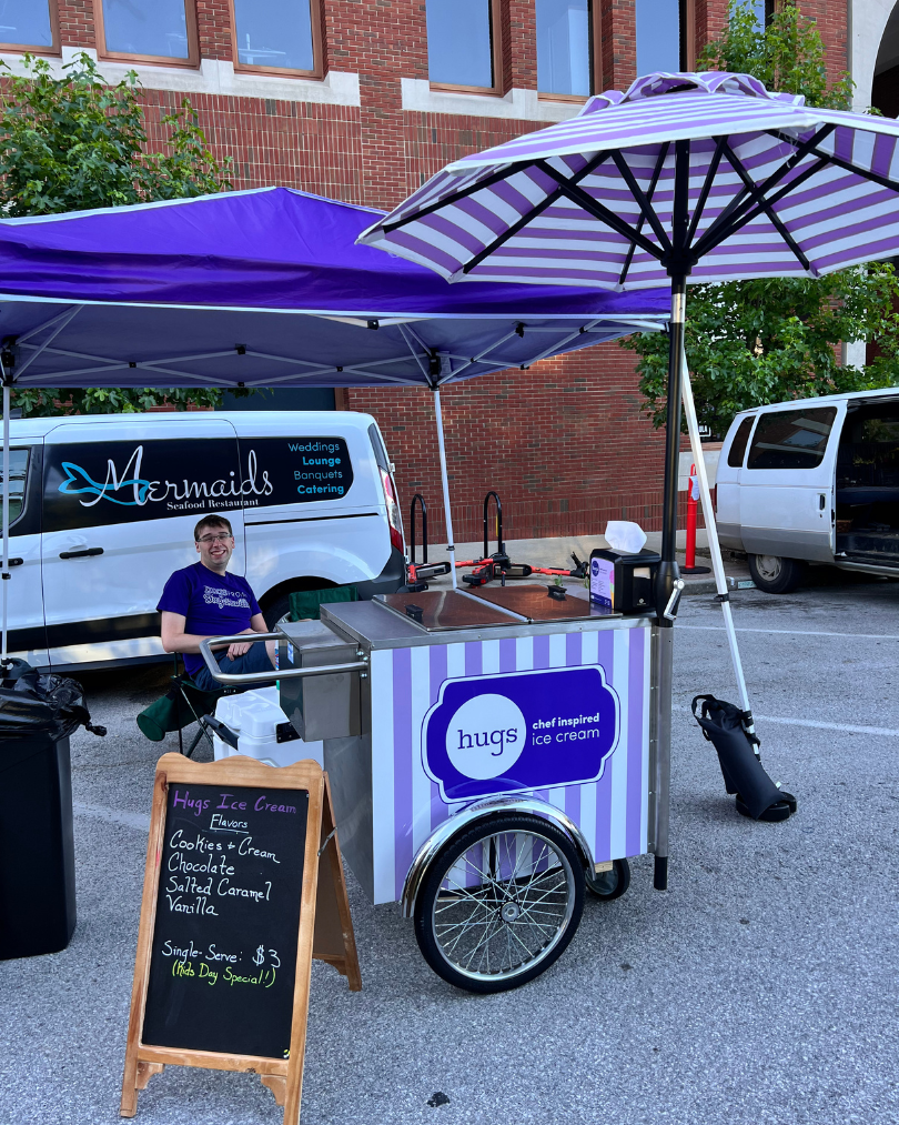 Hugs Ice Cream cart at a local event, featuring a smiling vendor, purple and white striped umbrella, and a menu board listing flavors like Cookies & Cream, Chocolate, Salted Caramel, and Vanilla.
