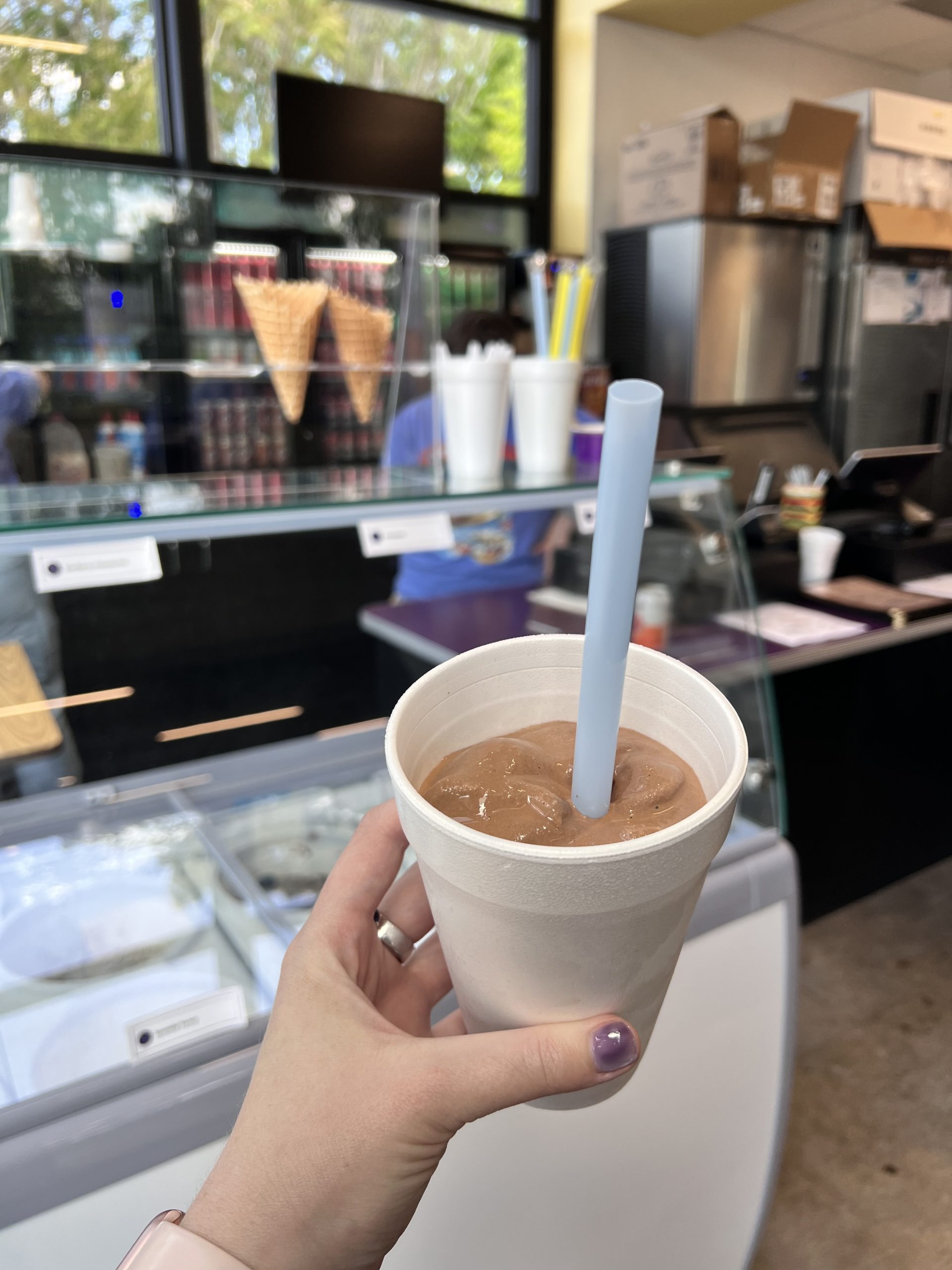 Hand holding a white cup with chocolate milkshake and a blue straw at an ice cream shop counter. behind-the-counter with cones and equipment visible